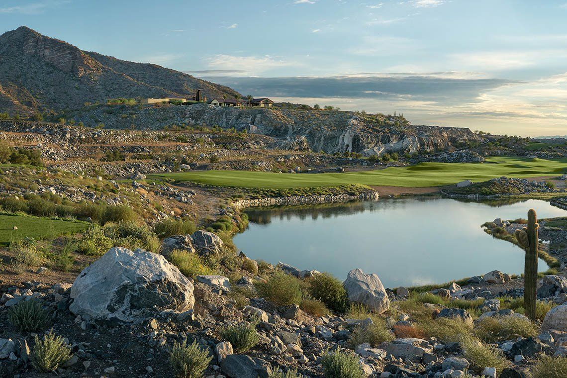 Scenic view of the 18th hole at Victory Golf Course in Victory at Verrado, Buckeye, Arizona, featuring rugged desert terrain, a serene water feature, and dramatic mountain landscapes, offering a challenging and picturesque golfing experience in this premier 55+ community.