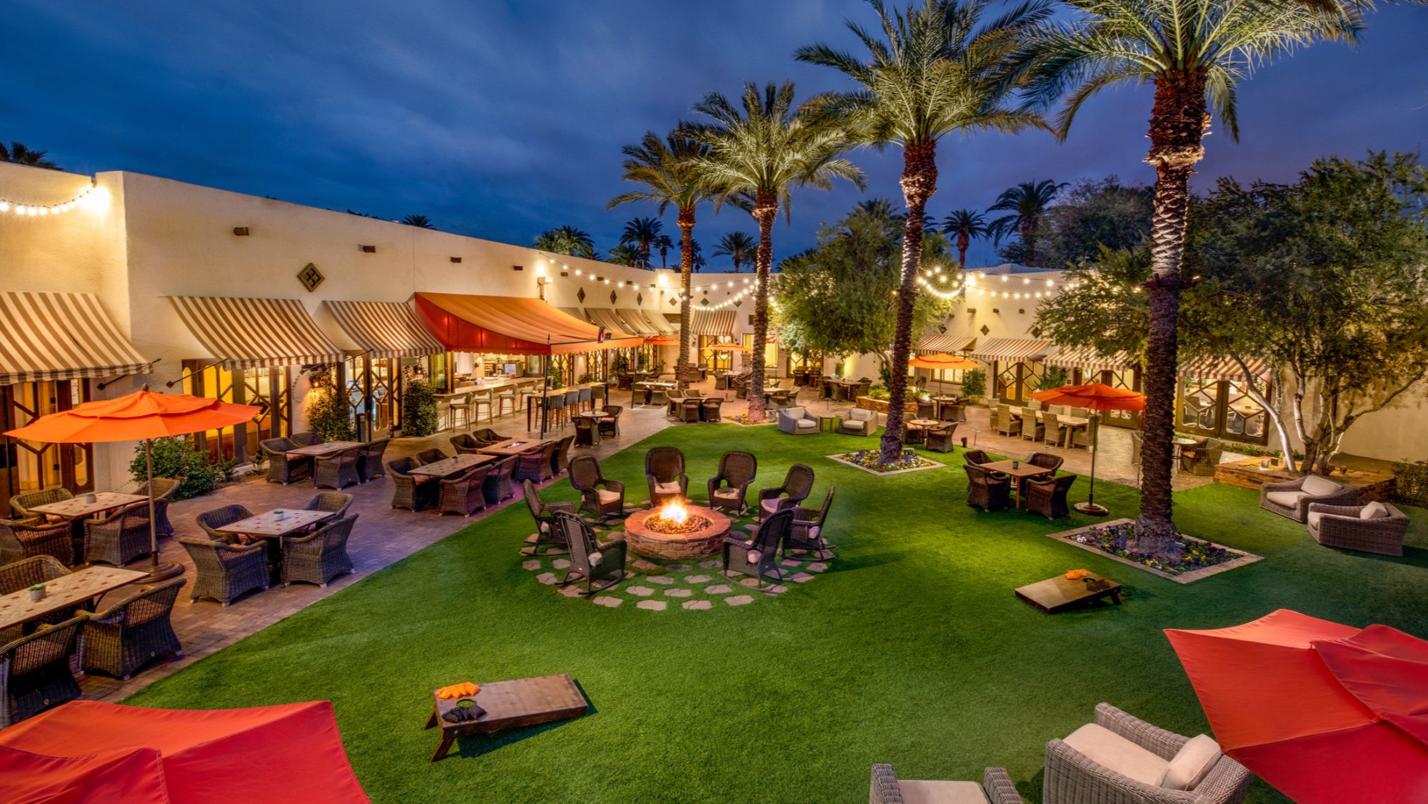 Outdoor dining and lounge area at the Wigwam Resort in Litchfield Park, AZ, illuminated with string lights and featuring fire pits, cozy seating, and palm trees. This luxurious Southwestern-style retreat offers a vibrant atmosphere for relaxation, dining, and socializing, making it a sought-after destination for residents and visitors alike.