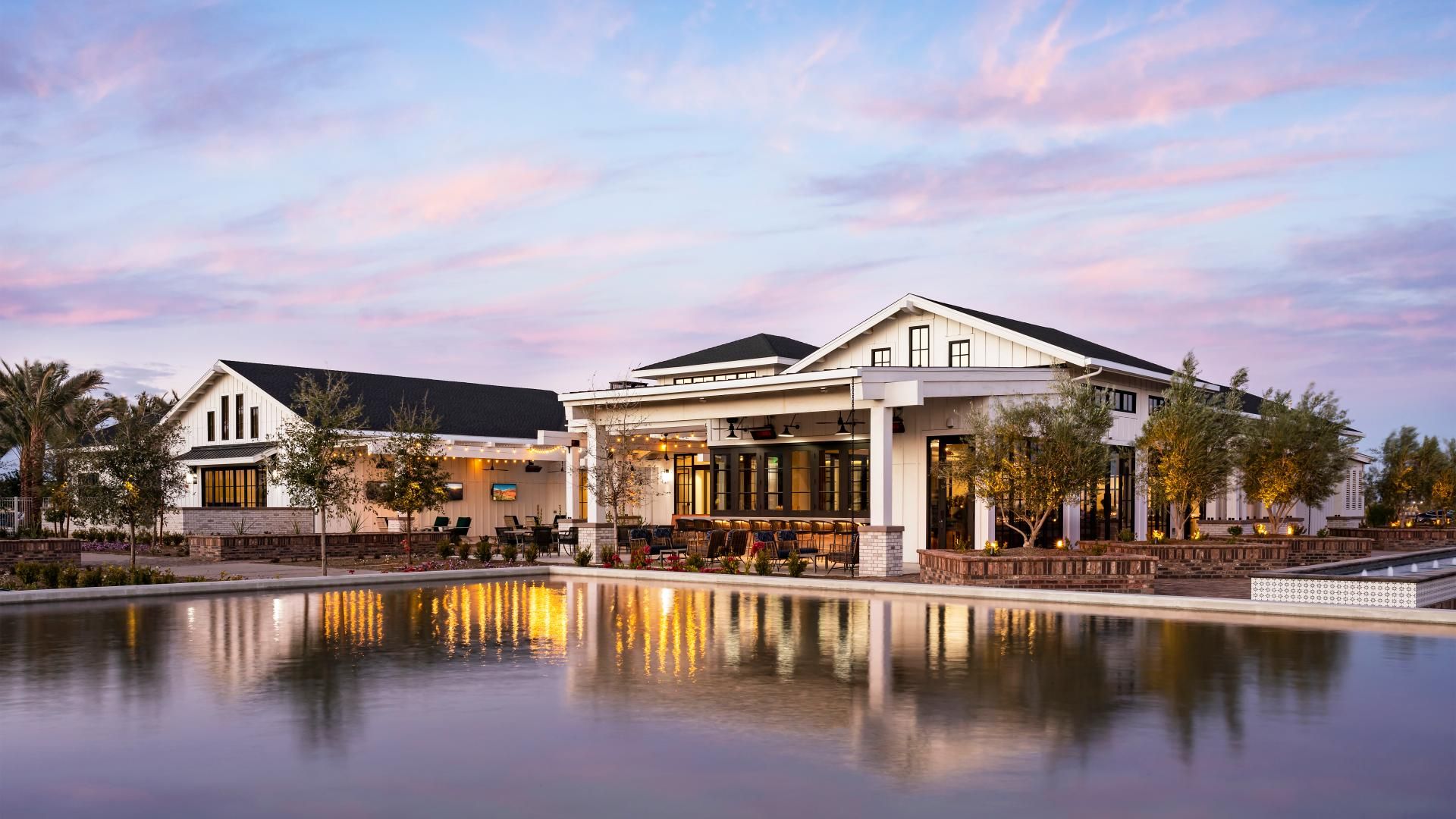 Elegant clubhouse at Sterling Grove in Surprise, Arizona, featuring modern farmhouse architecture, scenic waterfront views, and warm evening lighting. This luxurious clubhouse offers residents access to resort-style amenities including a golf course, fine dining, event spaces, fitness center, and a relaxing atmosphere with beautiful landscaping and mountain views. Ideal for those seeking an upscale lifestyle in a vibrant master-planned community surrounded by the beauty of the White Tank Mountains.