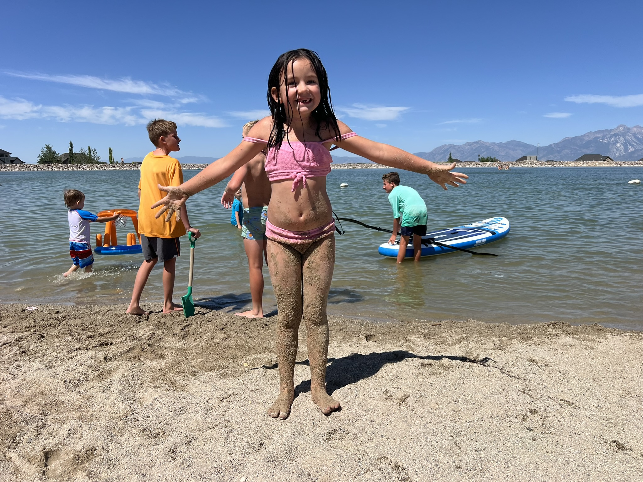 Quincy, a young girl in a pink swimsuit, smiling with open arms, covered in sand on the beach at Blackridge Reservoir. Other children play in the water and on the shore in the background, with paddleboards and floaties visible, all under a clear blue sky.