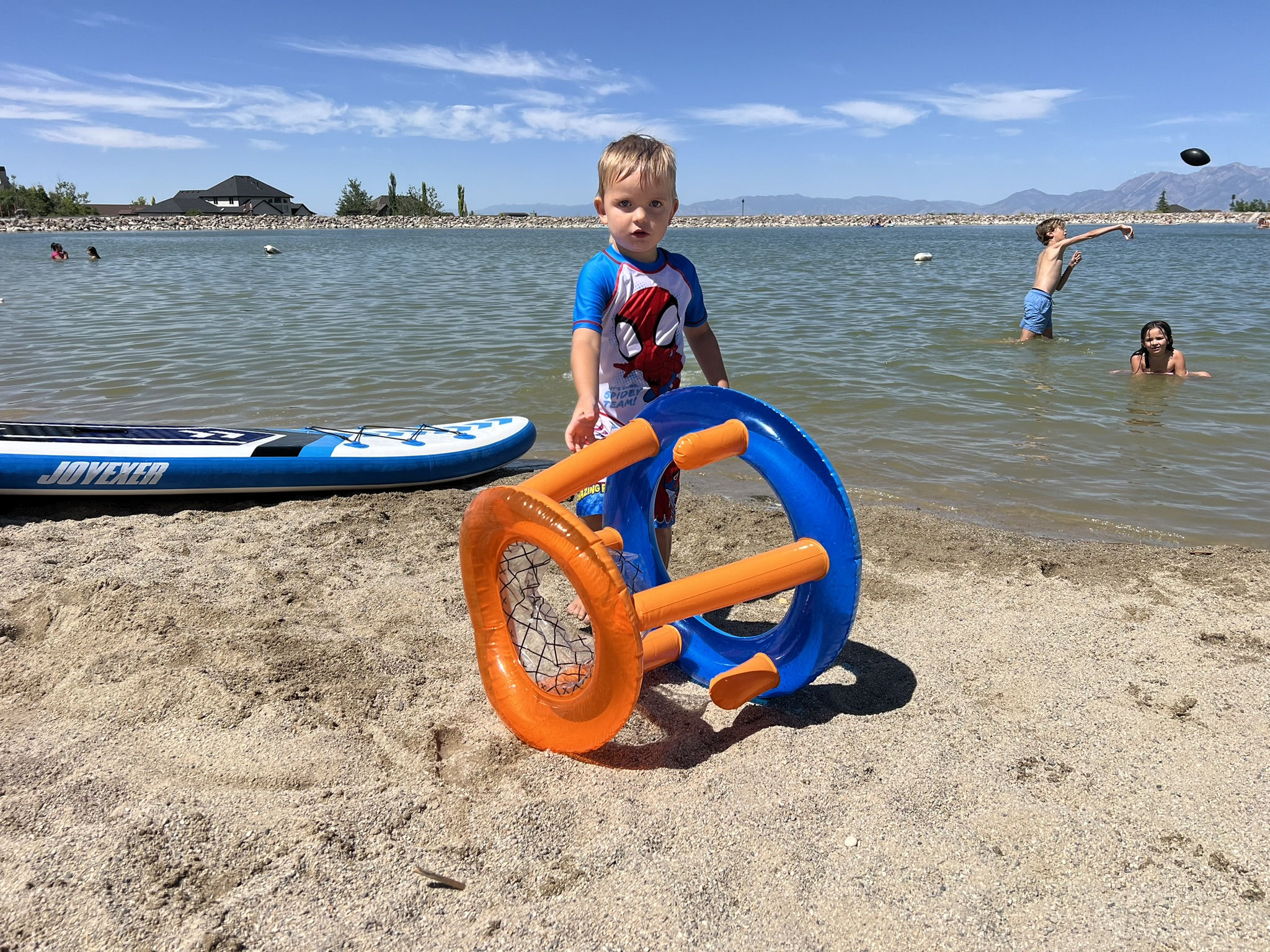 Trey, a young boy in a Spider-Man swimsuit, standing on the sandy beach at Blackridge Reservoir, holding an inflatable ring game. A paddleboard lies on the sand nearby, while other children play in the water in the background under a bright blue sky.