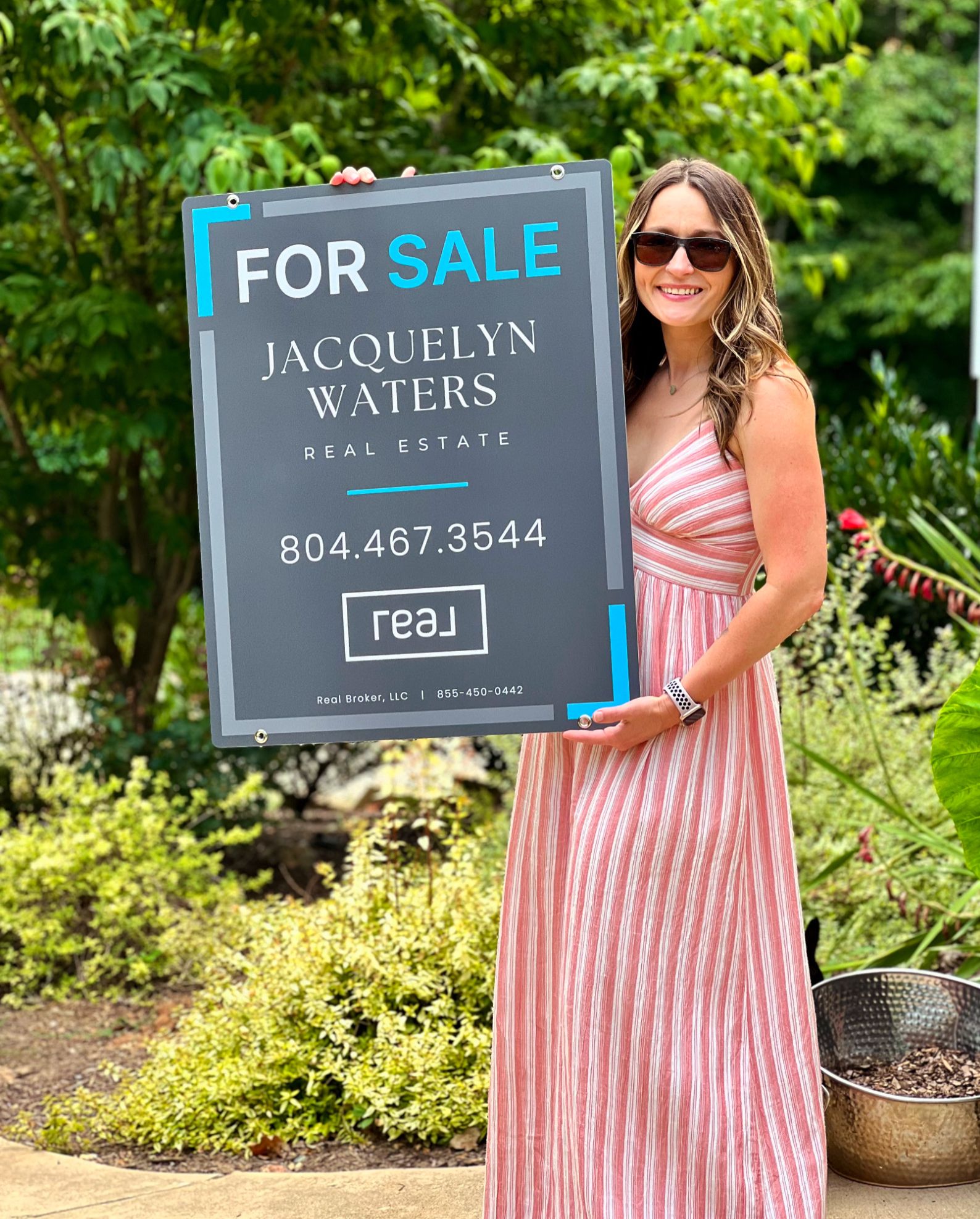 Jacquelyn Waters, Virginia Real Estate Agent, holding a 'For Sale' sign from Real Broker LLC, advertising properties for sale in Central Virginia.