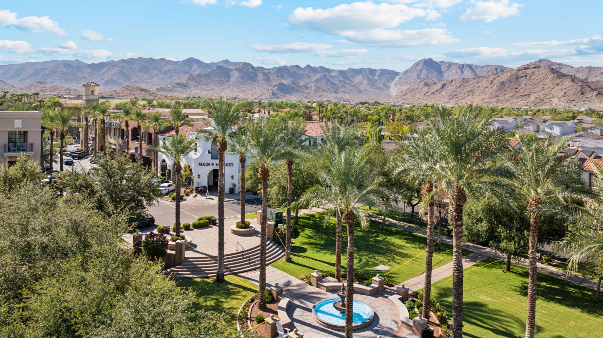 An aerial view of Verrado Downtown in Buckeye, Arizona, showcasing a vibrant community with tree-lined streets, a central park, charming shops, and restaurants. The area features a mix of residential homes with distinctive architecture, surrounded by lush greenery and mountainous desert landscapes in the background, highlighting the town's picturesque setting and walkable layout.