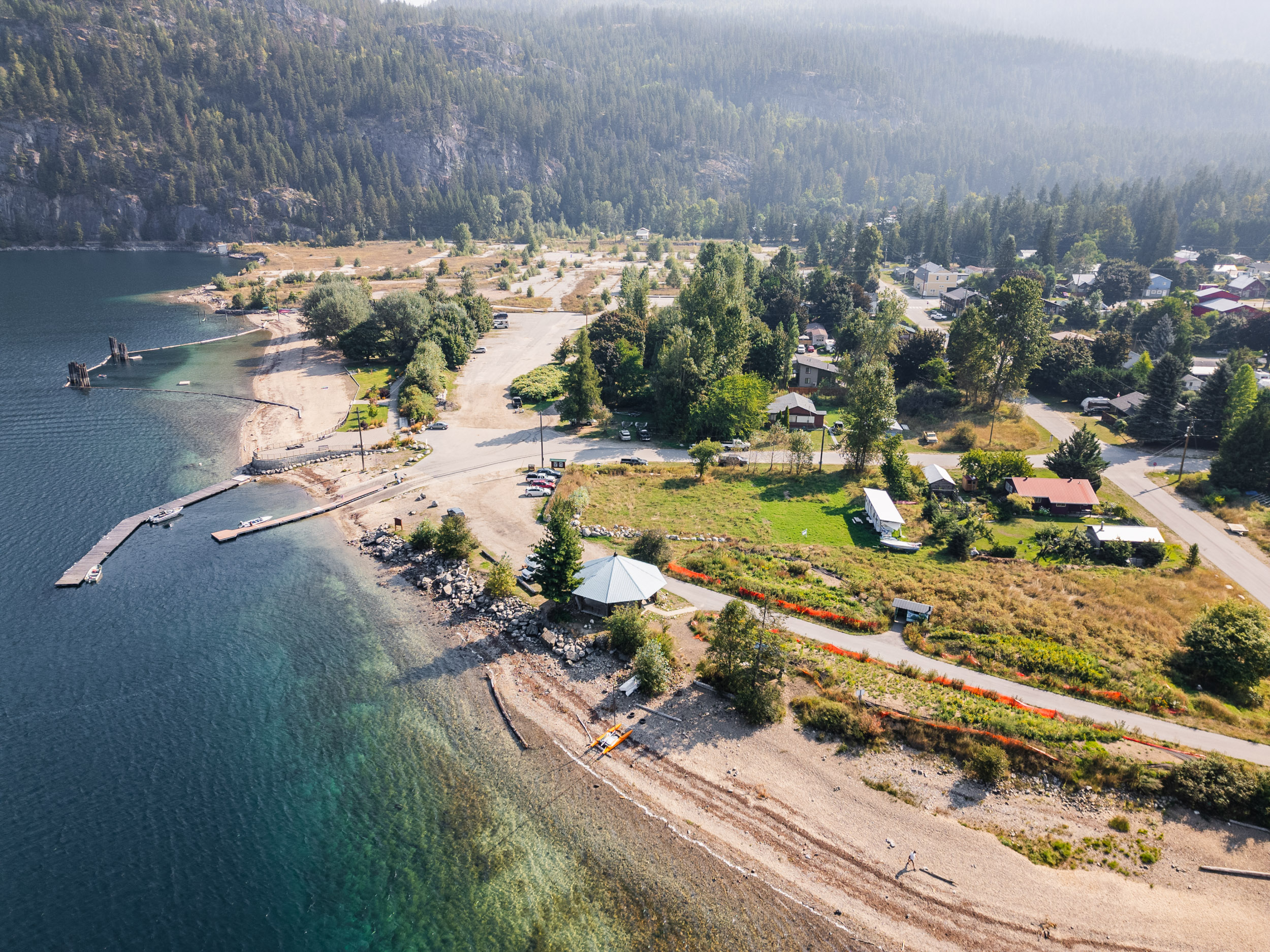 Aerial view of a lakeside community near a serene beach on Slocan Lake, surrounded by trees and mountains. The image shows homes, roads, and a dock extending into the calm waters of the lake