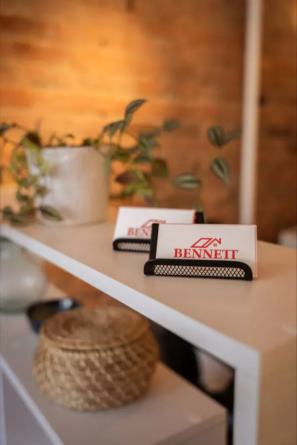 Business cards with the Bennett Family Real Estate logo displayed in a black metal holder on a white shelf, with a plant and decorative items in the background