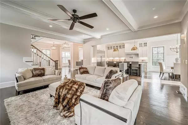 This image showcases a beautifully designed open-concept living space that flows seamlessly into the kitchen and dining areas. The living room features light, neutral-toned furniture, including two large sofas and two armchairs, arranged around a soft, plush area rug. The furniture is accented with luxurious fur throws and decorative pillows, adding warmth and texture to the space. A dark ceiling fan with sleek blades hangs above, contrasting with the room's bright and airy feel.  The kitchen is visible in the background, with white cabinetry and dark countertops. Two large pendant lights with metallic finishes hang above a central island with bar seating, providing a sophisticated and functional focal point. The dining area, located to the right, includes a modern dining table and chairs, bathed in natural light from the large windows.  Throughout the space, there are light wood floors and neutral walls, creating an elegant and inviting atmosphere. The overall design is modern, luxurious, and comfortable, perfect for both entertaining and everyday living.