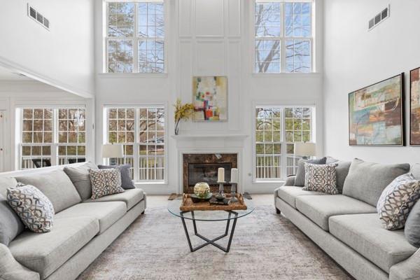 This image features a beautifully designed great room with high ceilings and large windows, allowing ample natural light to flood the space. The room is anchored by a central fireplace with a sleek, minimalist mantel, framed by tall windows that reach nearly to the ceiling, giving the room an airy and open feel. Above the fireplace is a piece of abstract art, adding a touch of color and modernity to the otherwise neutral decor. Two large, light gray sofas are symmetrically placed facing each other, creating a balanced and inviting seating area. The sofas are adorned with patterned throw pillows, adding texture and comfort to the setting. Between the sofas sits a contemporary glass coffee table with a geometric metal base, topped with decorative accents such as candles and a decorative sphere. The overall aesthetic is elegant and modern, with neutral tones and clean lines, while the height and openness of the room create a sense of grandeur and sophistication. This space is perfect for both entertaining guests and relaxing in a serene, well-lit environment.