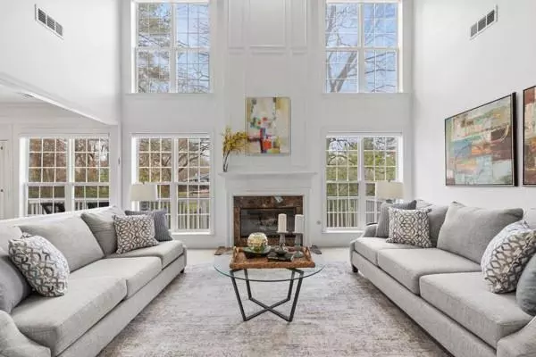 This image features a beautifully designed great room with high ceilings and large windows, allowing ample natural light to flood the space. The room is anchored by a central fireplace with a sleek, minimalist mantel, framed by tall windows that reach nearly to the ceiling, giving the room an airy and open feel. Above the fireplace is a piece of abstract art, adding a touch of color and modernity to the otherwise neutral decor.  Two large, light gray sofas are symmetrically placed facing each other, creating a balanced and inviting seating area. The sofas are adorned with patterned throw pillows, adding texture and comfort to the setting. Between the sofas sits a contemporary glass coffee table with a geometric metal base, topped with decorative accents such as candles and a decorative sphere.  The overall aesthetic is elegant and modern, with neutral tones and clean lines, while the height and openness of the room create a sense of grandeur and sophistication. This space is perfect for both entertaining guests and relaxing in a serene, well-lit environment.