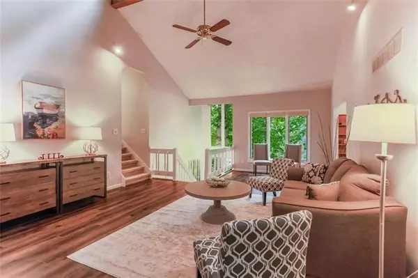 This image depicts a warm and inviting living room featuring a rustic design with a cozy, cabin-like atmosphere. The room's standout feature is its high, vaulted ceiling with exposed wooden beams and a rich, natural wood finish. The focal point of the room is a large stone fireplace with a wide hearth, creating a perfect space for gathering during colder months.   The furniture includes a comfortable brown sofa with decorative pillows, flanked by two patterned accent chairs, all arranged around a round, neutral-toned coffee table. The space is decorated with simple but tasteful elements, including a wrought iron wall piece and a floor lamp that casts soft lighting, adding to the room's welcoming ambiance.   The wooden flooring complements the ceiling's natural tones, enhancing the room's earthy, cozy feel. The room also features large windows that provide ample natural light, creating a balance between the warmth of the wood and the light, open atmosphere. The overall design exudes a comfortable, relaxed vibe, perfect for unwinding or entertaining guests.