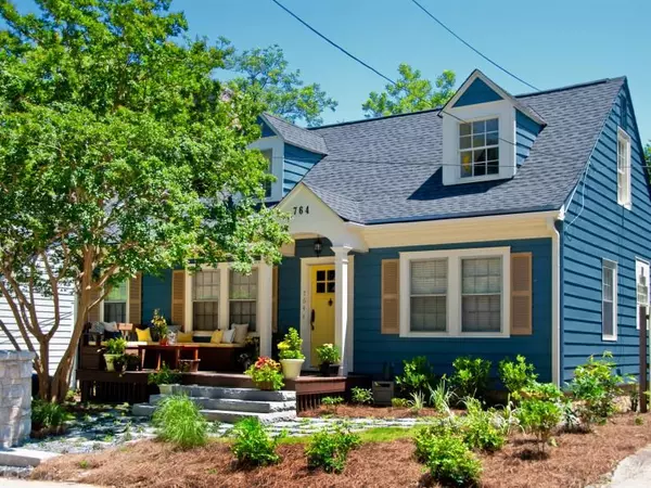 Charming blue cottage-style home with white trim, featuring a cozy front porch, landscaped garden, and welcoming yellow front door. The home includes dormer windows, a sloped roof, and a well-maintained yard with lush greenery.