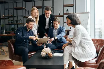 Group of professionals collaborating in a modern office setting, seated on chairs around a coffee table. They are engaged in a discussion, reviewing documents together in a casual, open workspace environment.