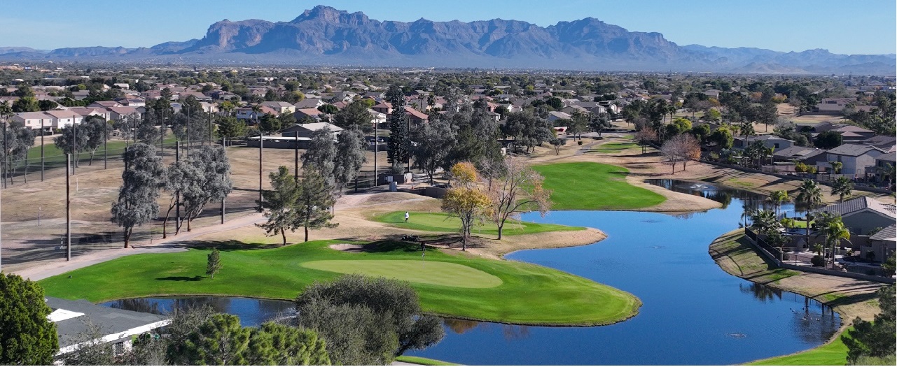 An aerial view of Augusta Ranch Golf Course in Mesa, AZ, showcasing the lush green fairways, neatly manicured landscaping, and picturesque desert surroundings. In the distance, the rugged silhouette of the Arizona mountains provides a stunning backdrop against a clear blue sky.