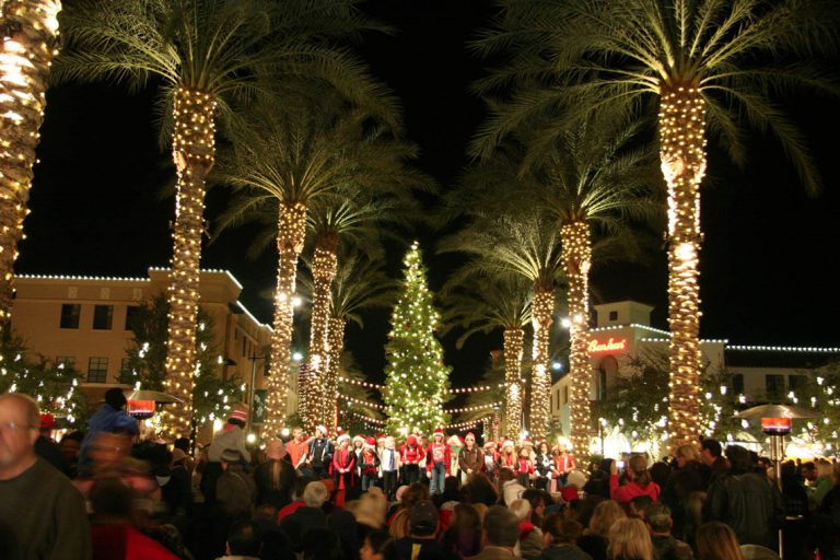 Nighttime view of downtown Verrado adorned with festive holiday lights, featuring a beautifully lit Christmas tree in the center. The buildings are outlined with twinkling lights, creating a warm and inviting holiday atmosphere under a clear night sky.