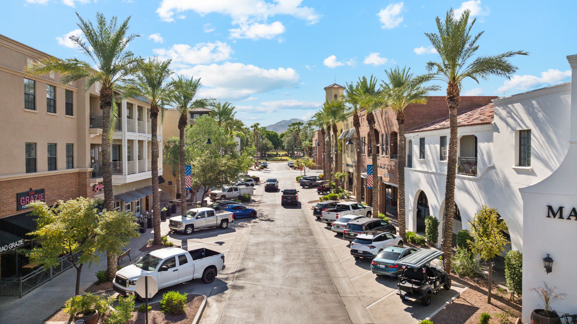 A picturesque view of downtown Verrado, featuring palm tree-lined streets with neatly manicured sidewalks. The area is bustling with charming storefronts and outdoor seating, capturing the vibrant and welcoming atmosphere of the community.