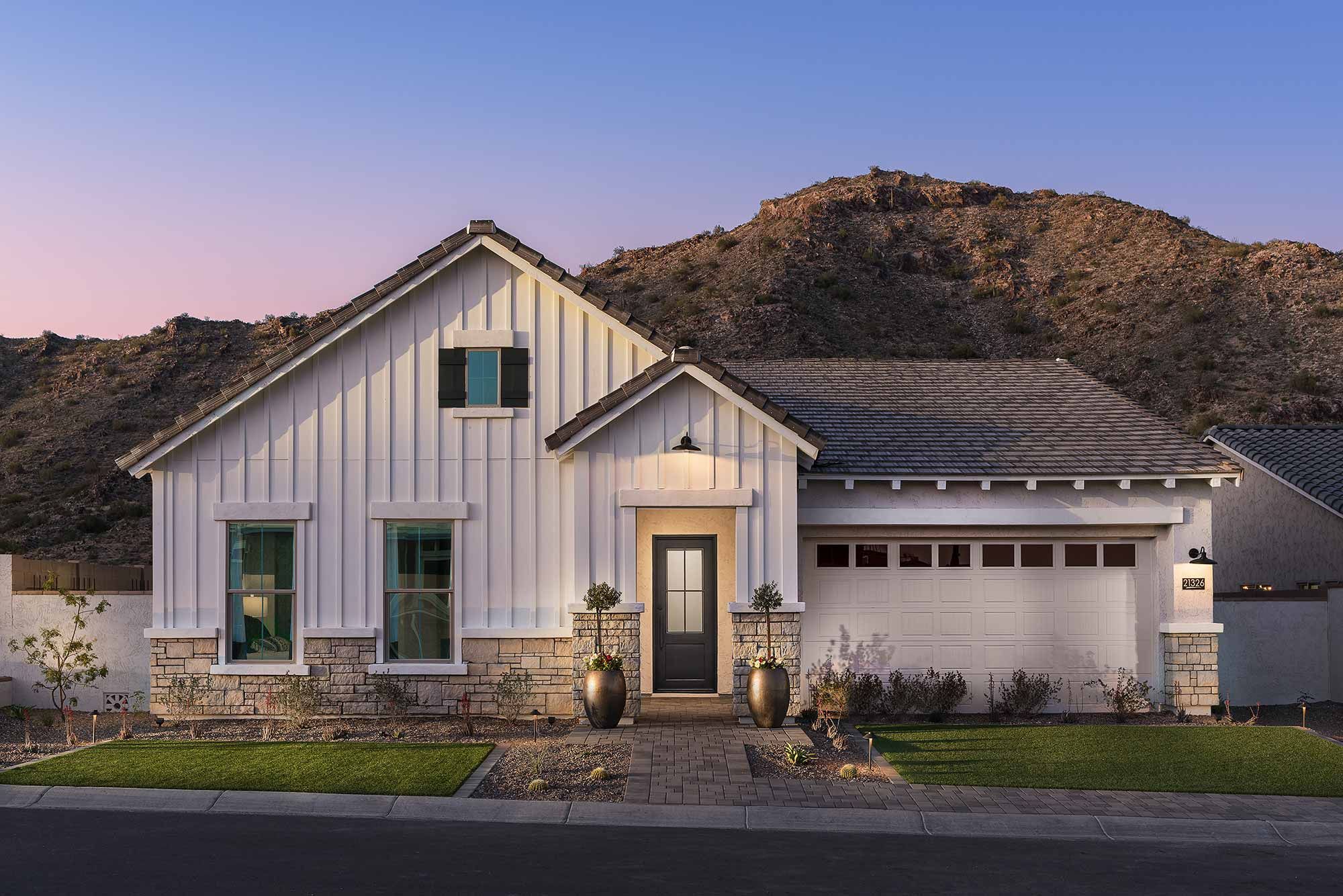 Newly constructed David Weekley home in the Verrado community, showcasing a modern, single-story design with a light beige exterior, large windows, and a beautifully landscaped front yard. The home features a spacious driveway leading to a two-car garage. In the background, stunning mountain views add a picturesque backdrop to the property, enhancing the serene and scenic atmosphere of the neighborhood.