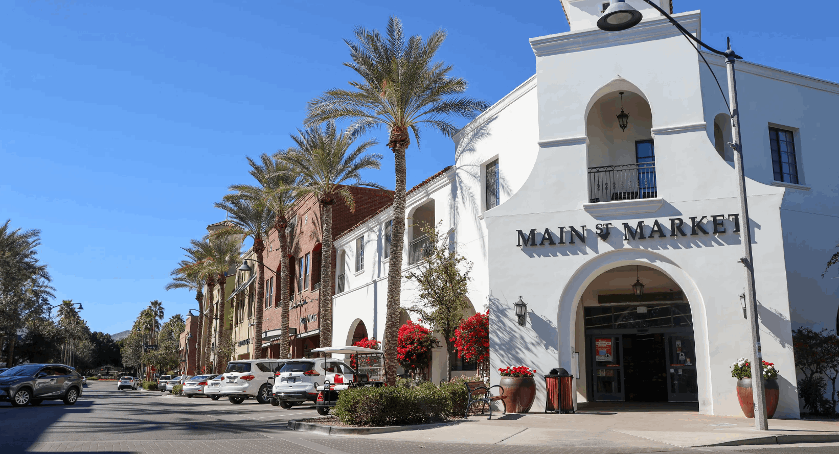 Vibrant Downtown Verrado in Buckeye, Arizona, featuring palm-lined streets, Spanish-style architecture, and the popular Main St Market under a bright blue sky. Cars are parked along the charming brick and stucco storefronts, creating a lively and inviting atmosphere.