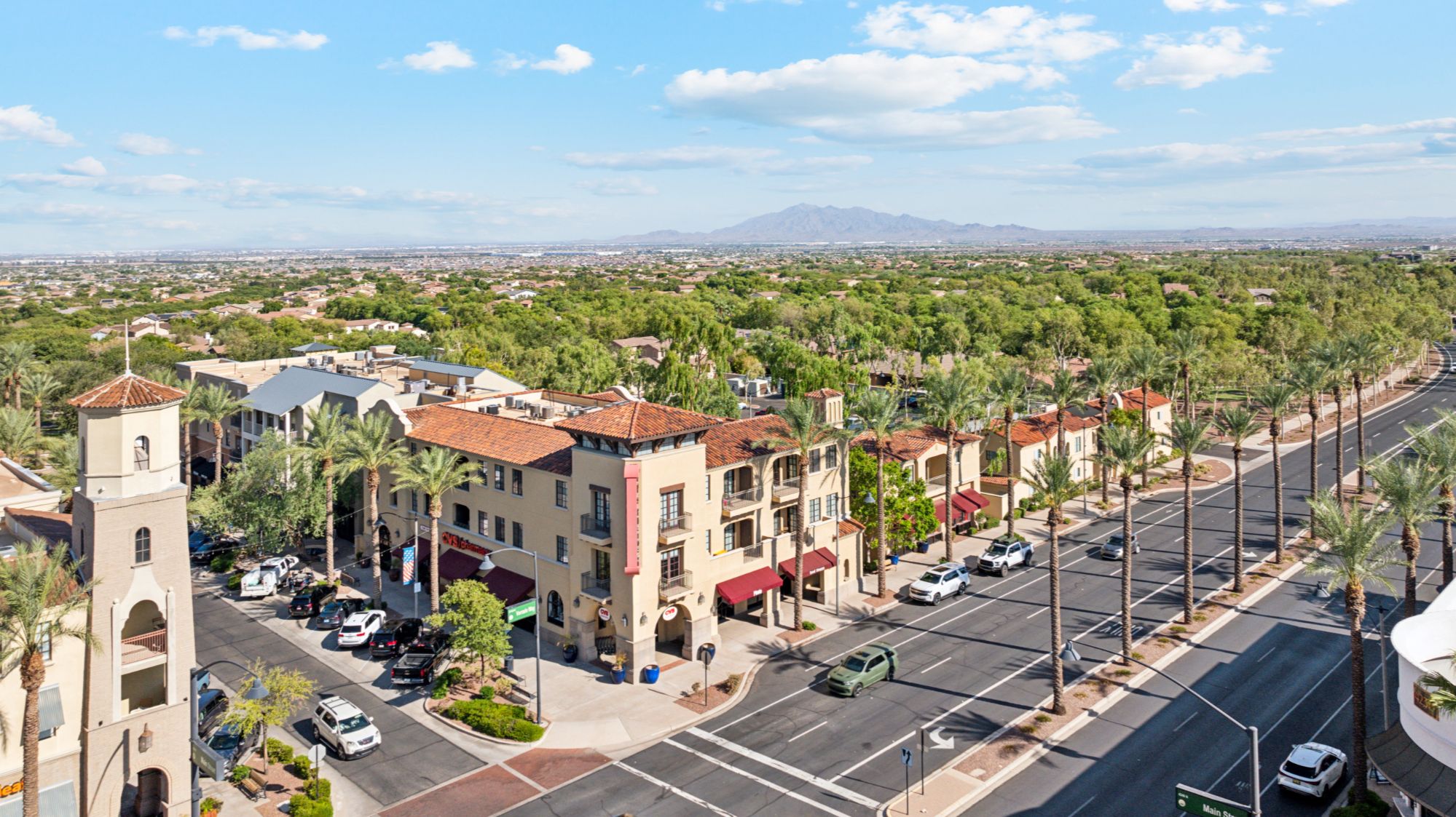 Arial shot of downtown Verrado, featuring a charming small-town atmosphere with tree-lined streets, neatly arranged buildings, and a central plaza. The surrounding area showcases a mix of residential homes with rooftops visible, set against a backdrop of rolling hills and a desert landscape, highlighting Verrado's blend of natural beauty and community charm