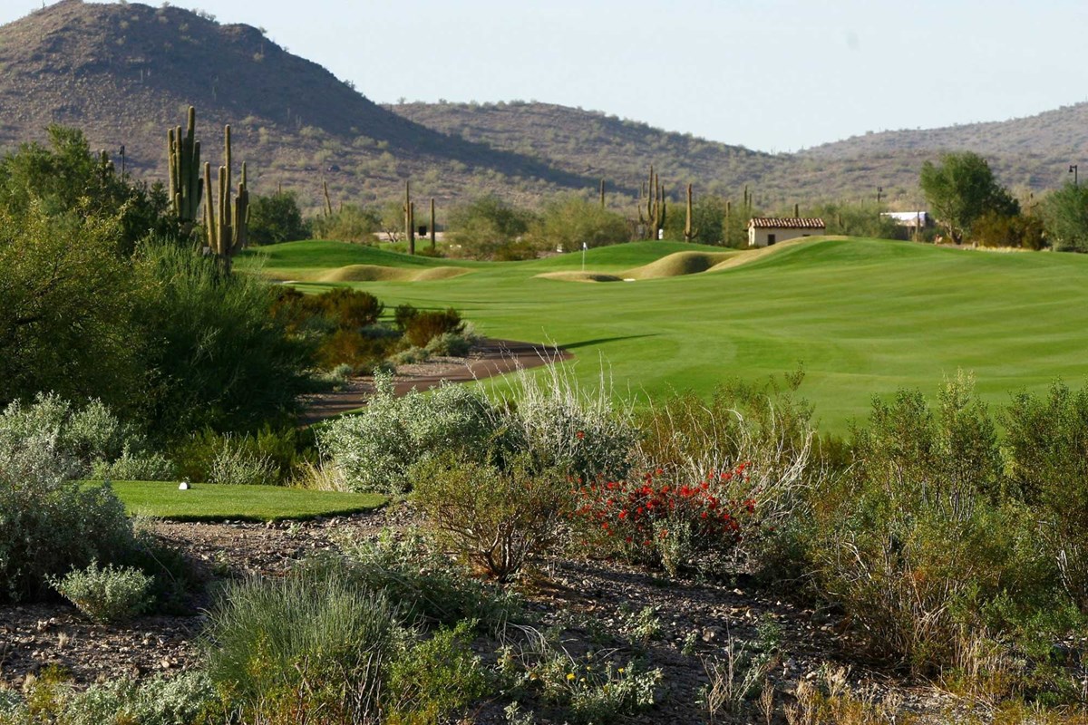 Scenic view of a lush green golf course in the Vistancia community of Peoria, AZ, with rolling fairways, strategically placed bunkers, and a backdrop of desert mountains under a clear blue sky.