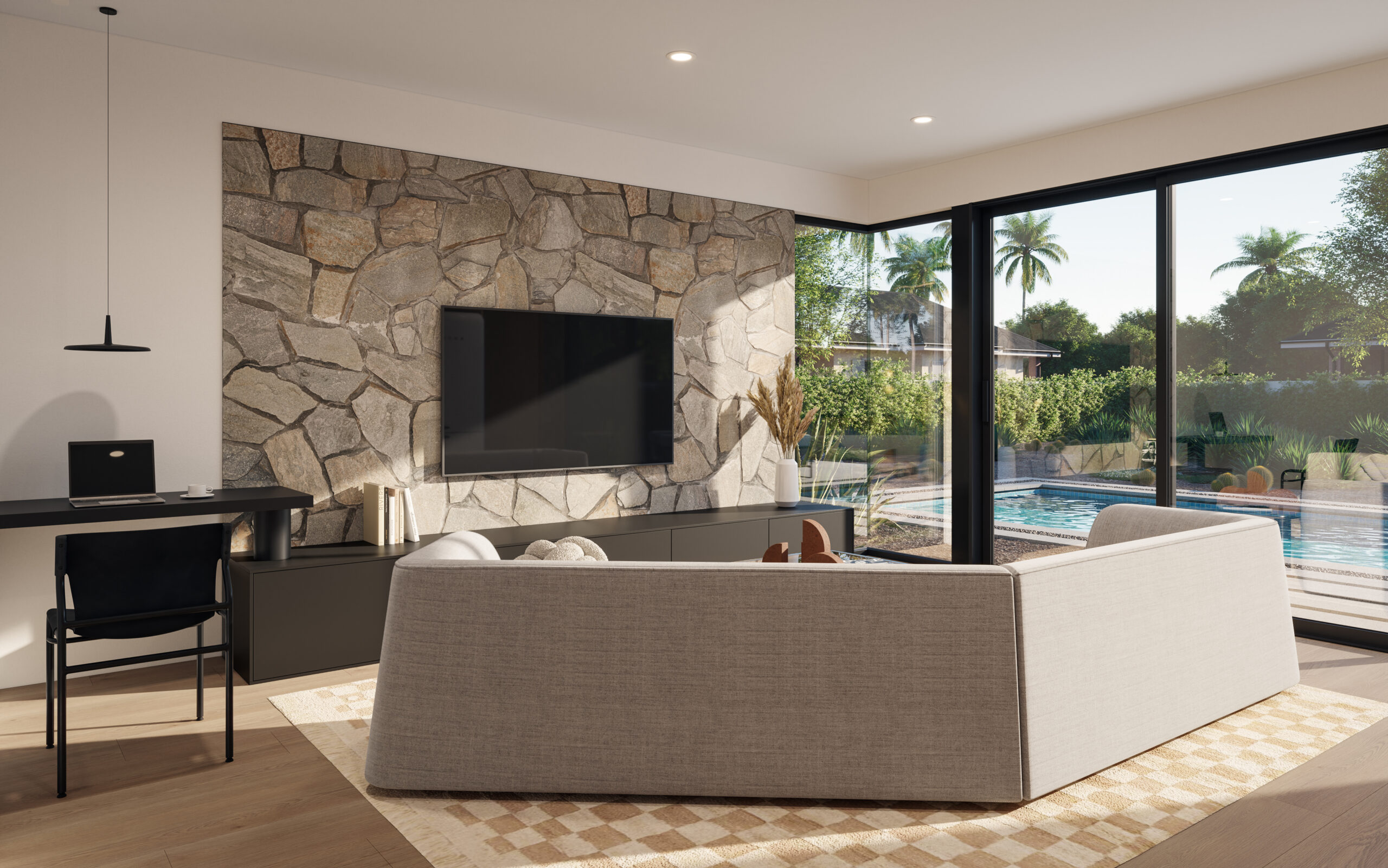 Modern living room in an Arizona ADU featuring a cozy beige sofa, a stone accent wall with a mounted flat-screen TV, and large floor-to-ceiling sliding glass doors providing a view of a pool and desert landscaping with palm trees. The space is decorated with minimalist furniture, including a black desk with a laptop, pendant lighting, and neutral tones, creating a serene and inviting atmosphere that blends indoor and outdoor living.