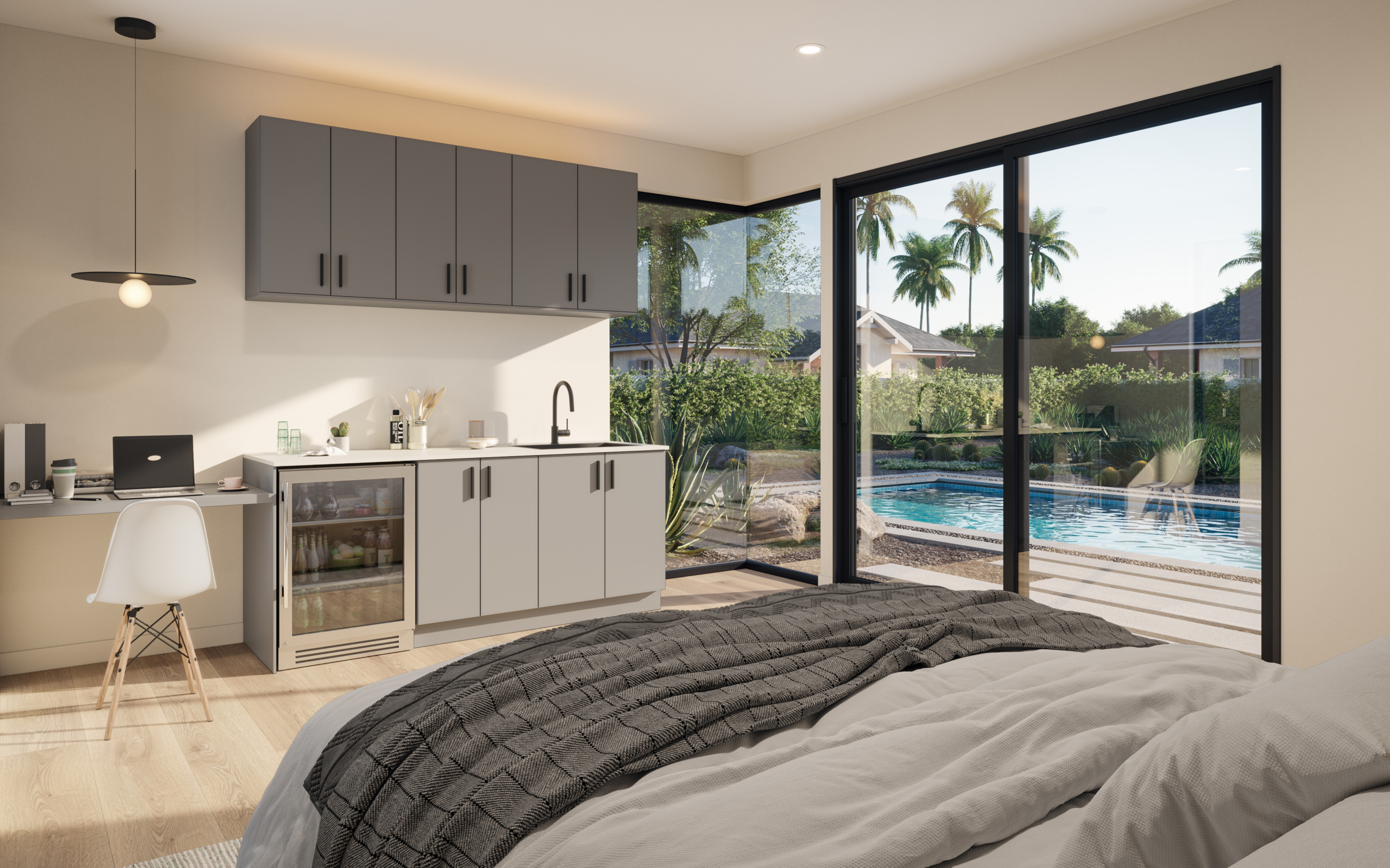 Interior of a modern bedroom in an Accessory Dwelling Unit (ADU) in Arizona. The room features a minimalist design with light wood flooring, a cozy bed covered in neutral bedding, and a small kitchenette with grey cabinets, a sink, and a mini-fridge. A desk with a laptop is positioned under a stylish pendant light. Large sliding glass doors provide a view of an outdoor pool surrounded by desert landscaping, including palm trees and greenery, creating a serene atmosphere.