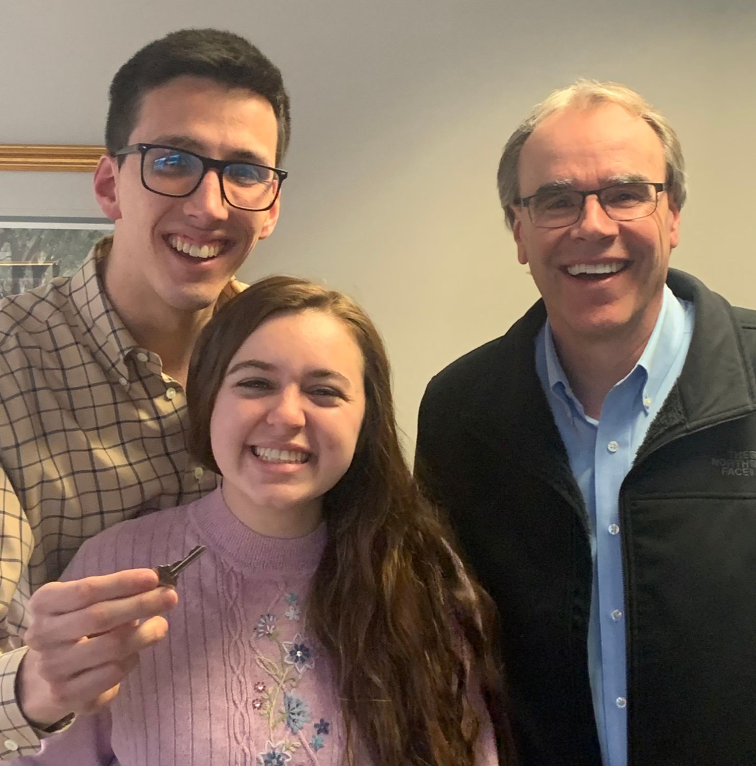 Two happy clients - a man and woman - stand next to David Campbell, Realtor - all smiling and facing towards the camera. The man is holding a house key.