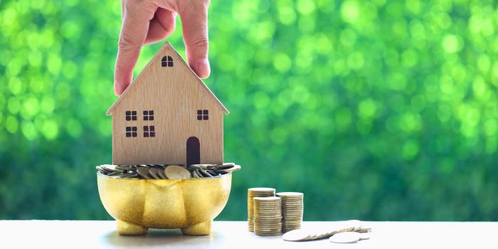 A hand placing a wooden house model into a golden bowl filled with coins, symbolizing real estate as a powerful wealth-building investment. Stacks of coins on the side represent financial growth and stability in 2025.