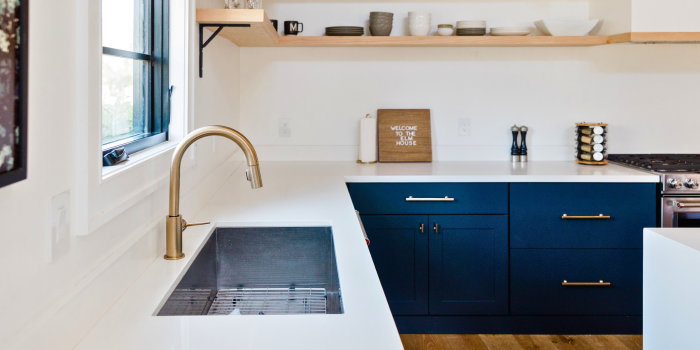 A modern farmhouse kitchen featuring deep navy cabinetry with gold hardware, open wood shelving with minimalist decor, and a sleek white countertop paired with a gold faucet, blending traditional charm with contemporary elegance.