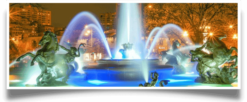 Nighttime fountain with illuminated water jets and bronze sculptures of horses and mythical figures, set against a backdrop of city buildings.