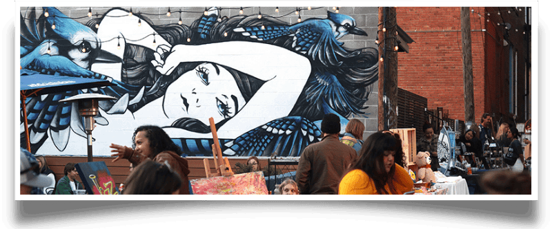 Outdoor scene with a large mural of a woman and blue jays, people relaxing and browsing at a street fair.