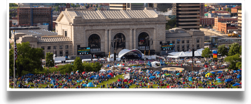 Large crowd gathered in a park-like area in front of a historic train station, with tents and stage setup for an outdoor event.