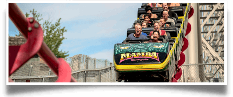 People on a yellow and green Mamba roller coaster train, ascending a hill at a theme park.