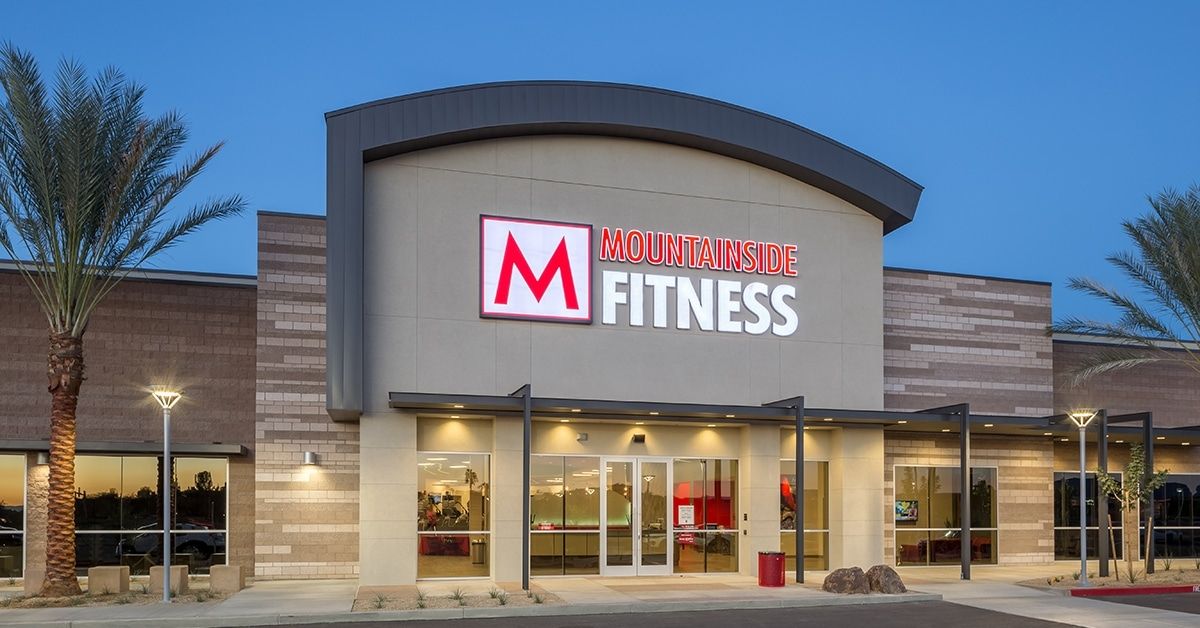 Exterior of the upcoming Mountainside Fitness in Vistancia, Peoria, Arizona, featuring a modern design with large glass windows, palm trees, and well-lit entryway. This state-of-the-art gym will provide premium fitness amenities, including strength training, cardio equipment, group classes, personal training, and luxury locker rooms. Strategically located to serve the growing Vistancia community, Mountainside Fitness will enhance local health and wellness options, making it a top fitness destination in the area.