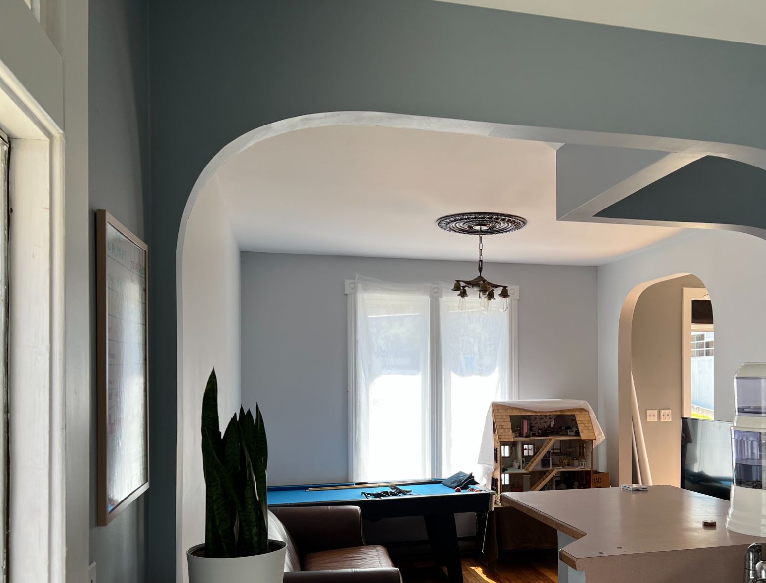 A cozy living space with arched entryways and soft blue-gray walls. The image features a small kitchen counter with a double sink and a water filter, adjacent to a living area with natural light streaming through sheer white curtains. A brown leather couch sits beside a blue pool table, and a dollhouse is visible in the corner. The hardwood flooring adds warmth to the space, while a potted plant and a whiteboard with markers bring functional decor elements.