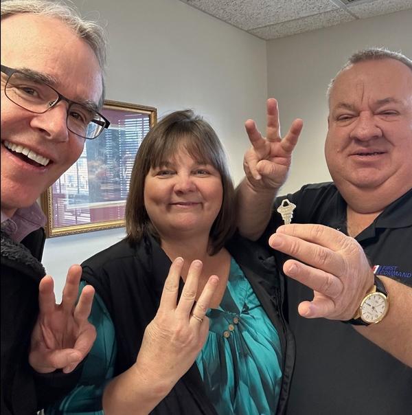 David Campbell stands besides a man and woman - all are smiling and are holding up three fingers. The man is holding up a key.